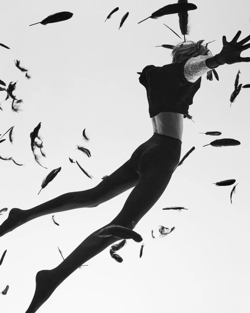 Black-and-white studio shot of a slender person jumping amidst floating feathers against a bright background, captured with a low-angle view.