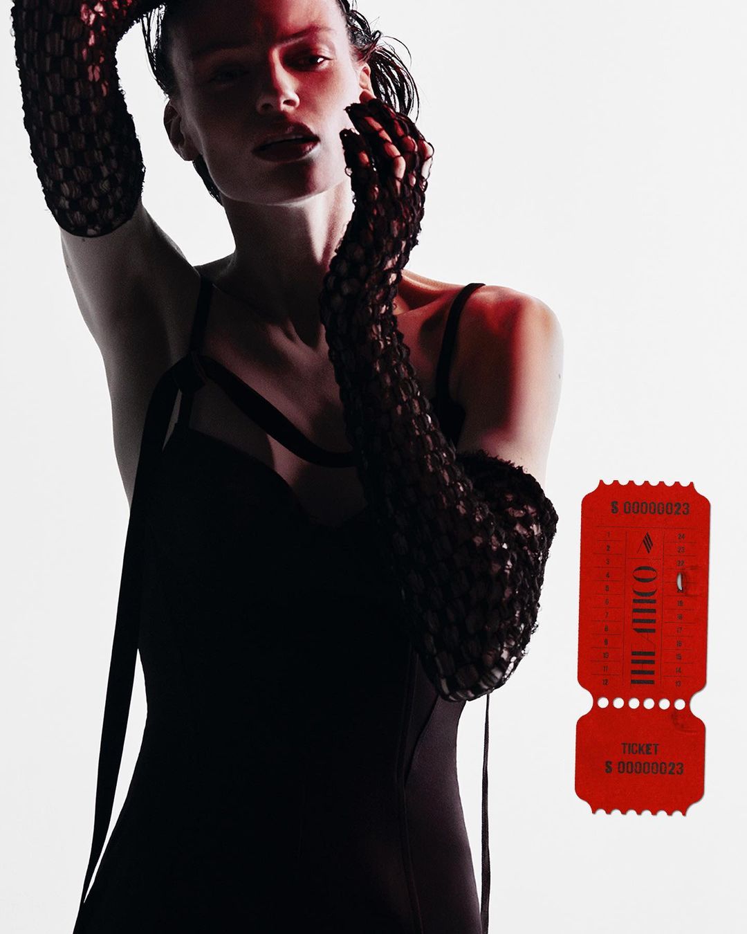 A female fashion model poses in a black dress and arm-length mesh gloves in a studio with a red ticket prop and bright lighting.