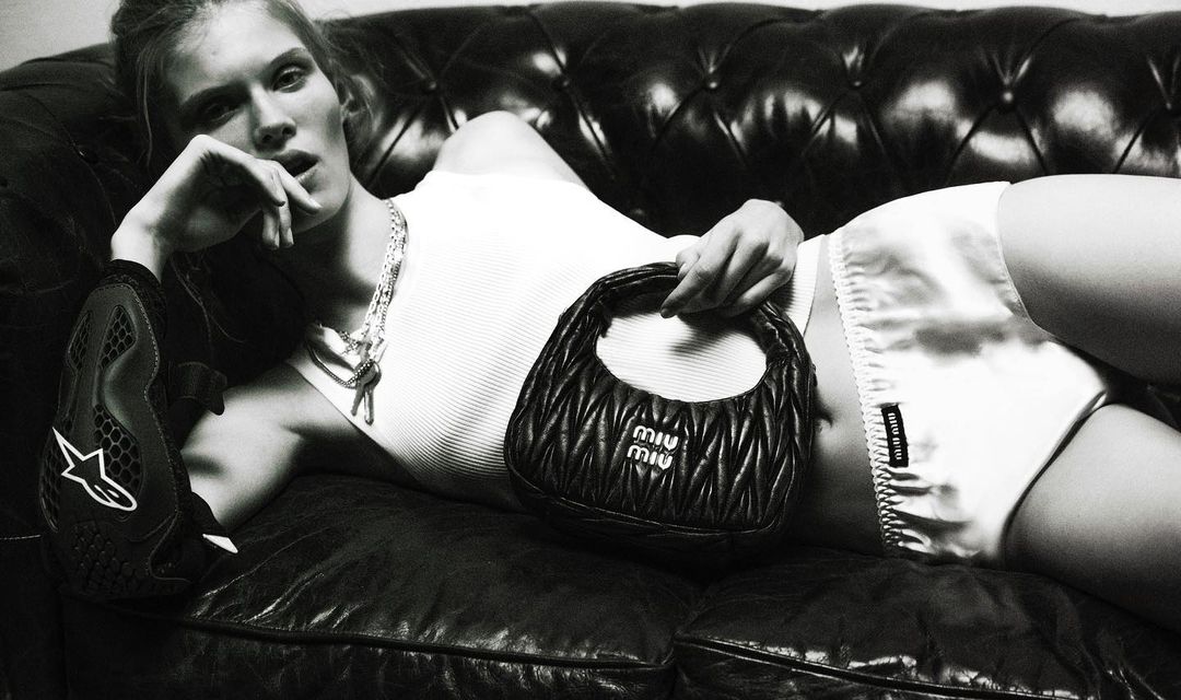 Monochromatic studio shot of a blonde woman reclining on a leather couch while holding a black Miu Miu handbag.