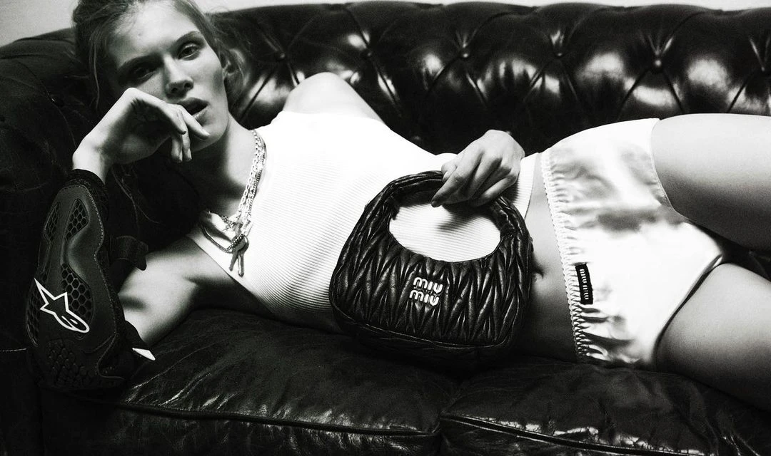 Monochromatic studio shot of a blonde woman reclining on a leather couch while holding a black Miu Miu handbag.