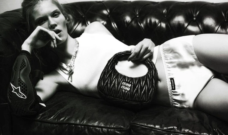 Monochromatic studio shot of a blonde woman reclining on a leather couch while holding a black Miu Miu handbag.