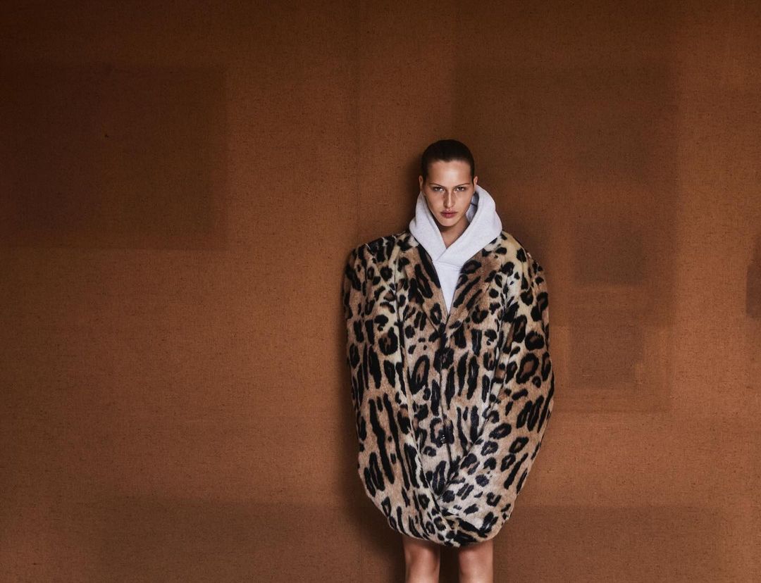 Woman posing in a white hoodie and a leopard-print faux fur coat against a brown studio backdrop, fashion editorial shot.