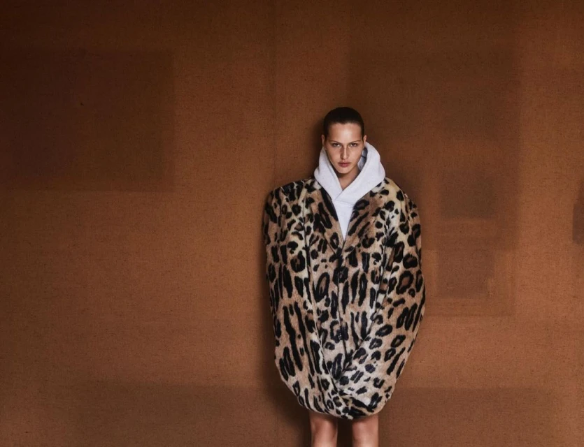 Woman posing in a white hoodie and a leopard-print faux fur coat against a brown studio backdrop, fashion editorial shot.