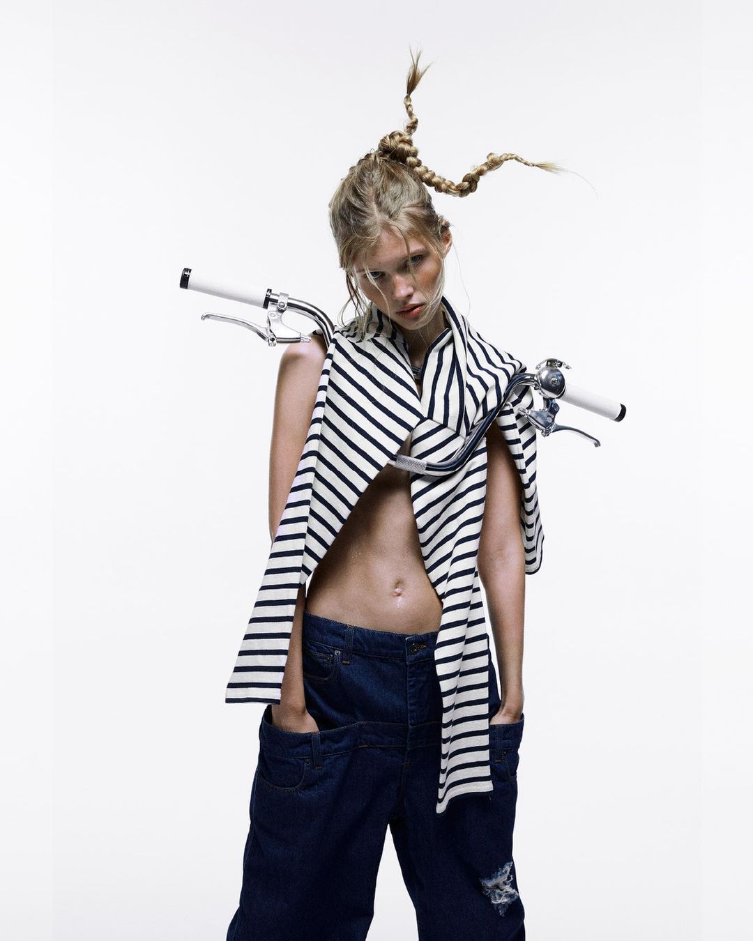Fashion shot of a woman with a unique braided hairstyle, wearing a striped shirt and denim pants, with bicycle handlebars around her neck.