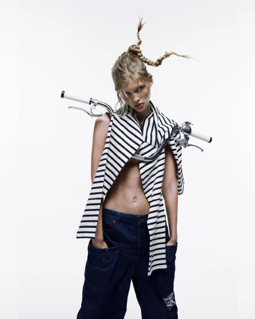 Fashion shot of a woman with a unique braided hairstyle, wearing a striped shirt and denim pants, with bicycle handlebars around her neck.