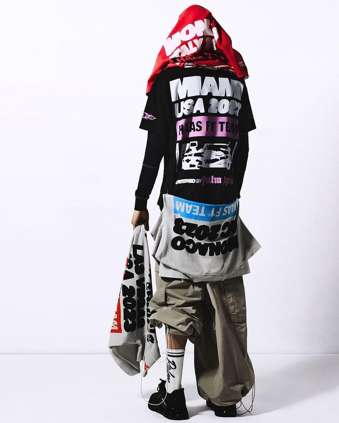 Person in oversized streetwear fashion stands against a white backdrop wearing racing-themed graphic tees and baggy cargo pants.