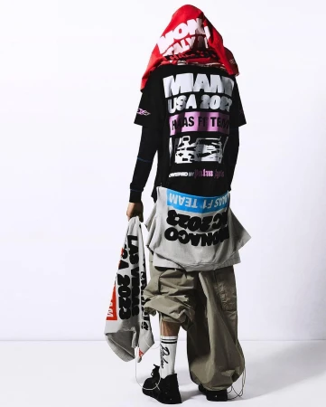 Person in oversized streetwear fashion stands against a white backdrop wearing racing-themed graphic tees and baggy cargo pants.