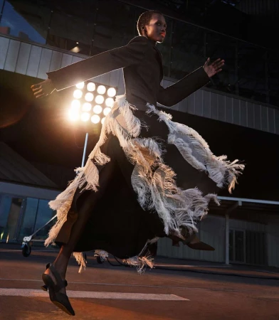 Model in a black blazer and fringe skirt mid-air with a bright spotlight in the background, photographed by Vito Fernicola.