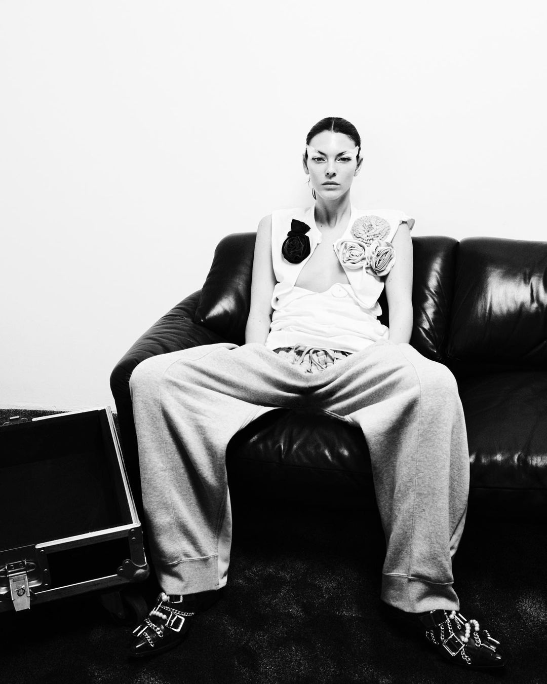 Black and white portrait of adult female model sitting on black couch with open metal suitcase, wearing white top and sweatpants.