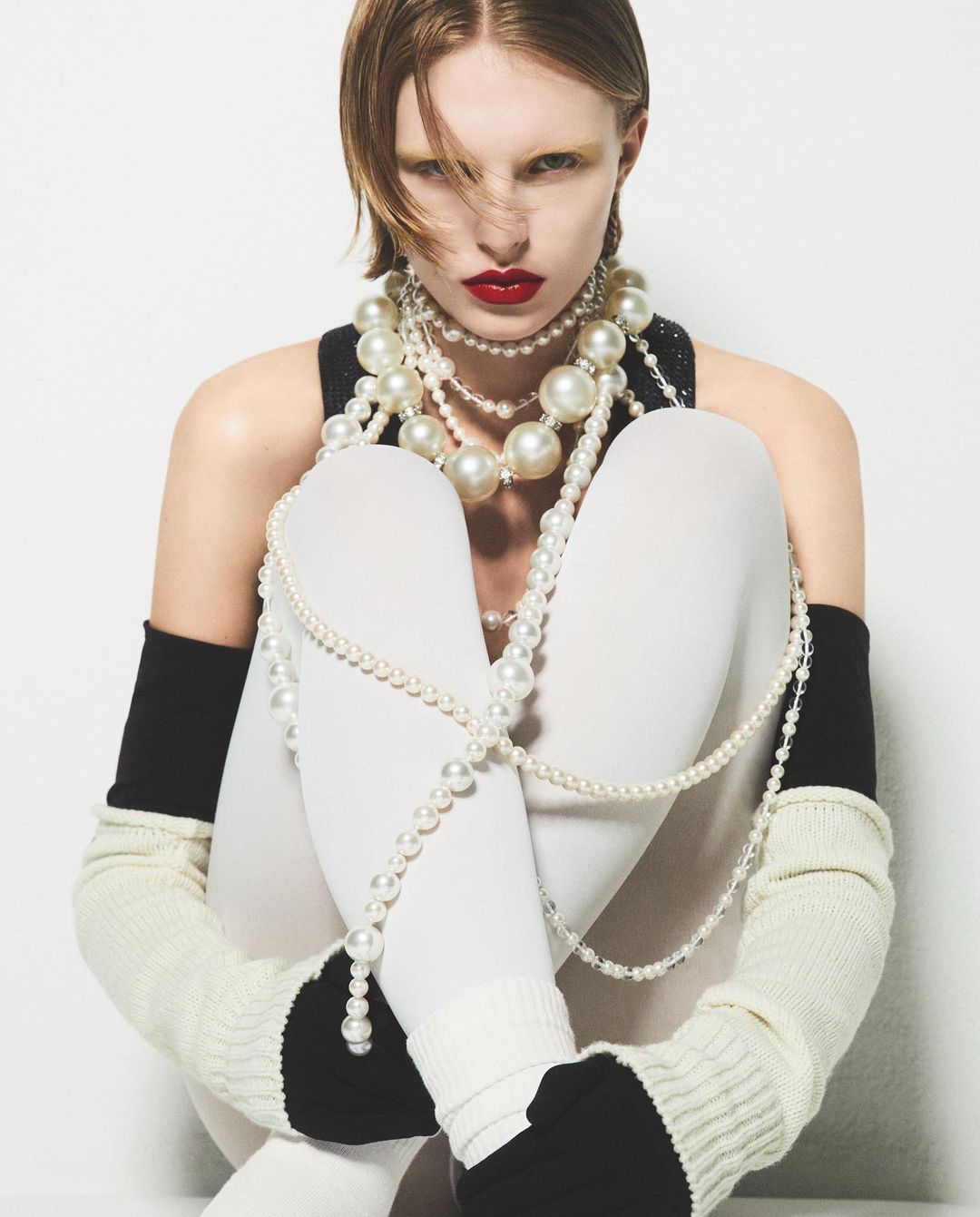 Fashion portrait of a pale-skinned woman with red lips wearing a black top and white tights, adorned with multiple strands of pearls.