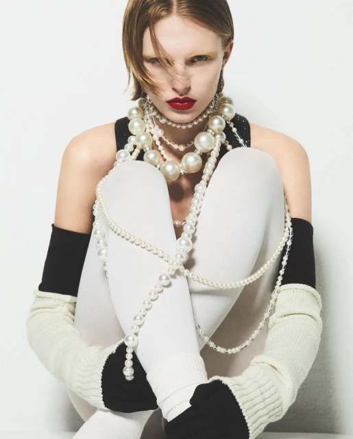 Fashion portrait of a pale-skinned woman with red lips wearing a black top and white tights, adorned with multiple strands of pearls.
