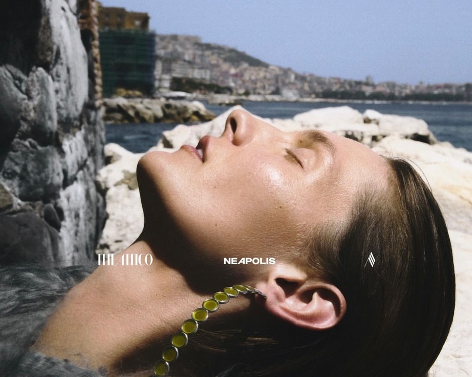 Woman with necklace reclines by the sea against a rocky backdrop in a summery outdoor scene with distant city views.
