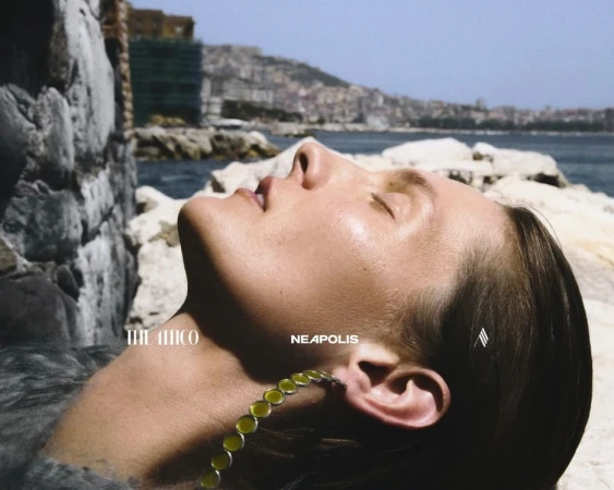 Woman with necklace reclines by the sea against a rocky backdrop in a summery outdoor scene with distant city views.