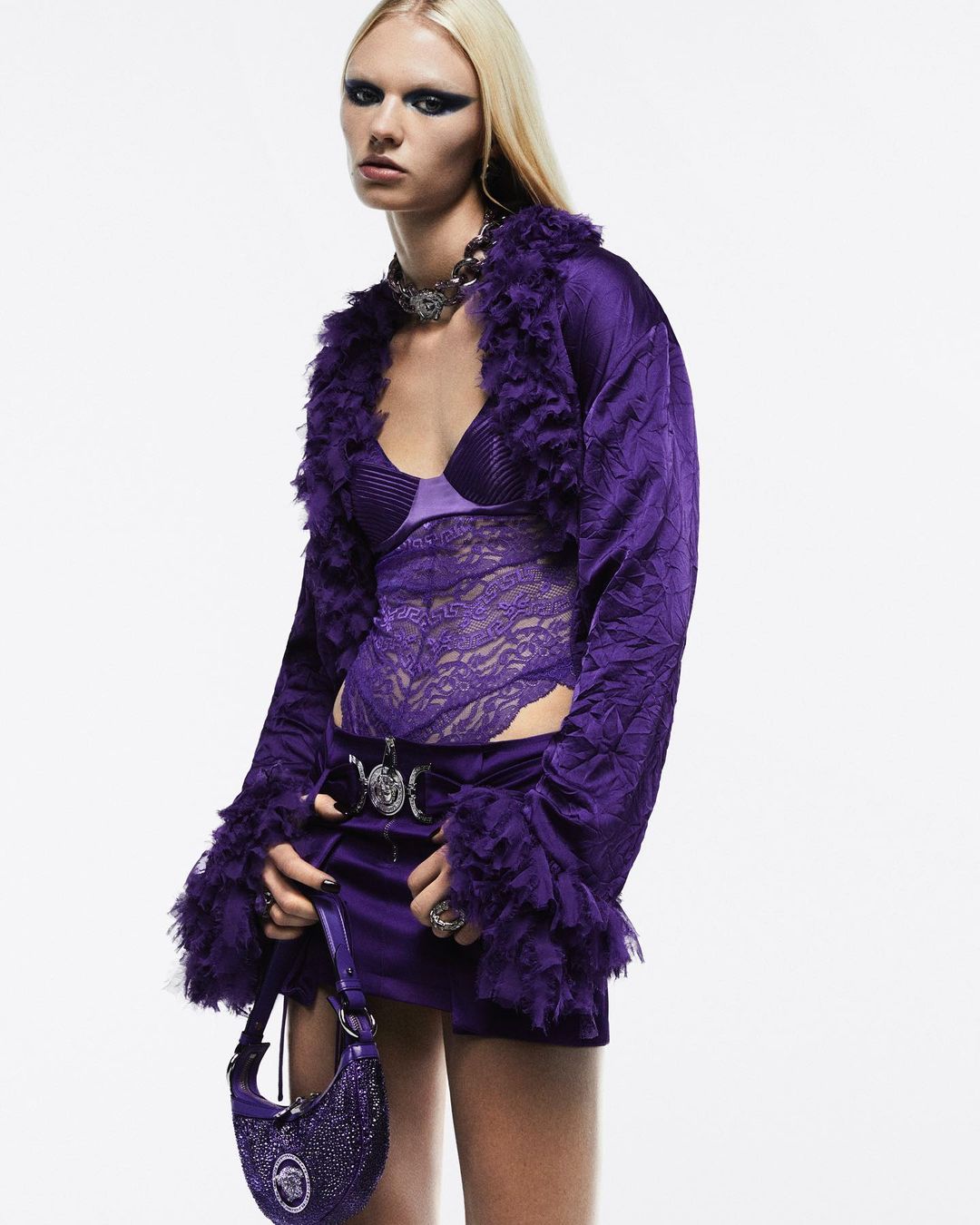 Fashion shot of a woman wearing a purple jacket with fur trim and a matching skirt and purse against a white backdrop.