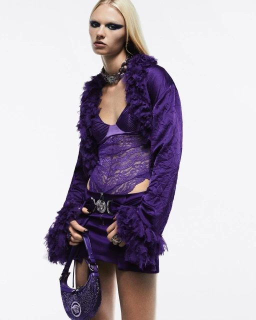 Fashion shot of a woman wearing a purple jacket with fur trim and a matching skirt and purse against a white backdrop.