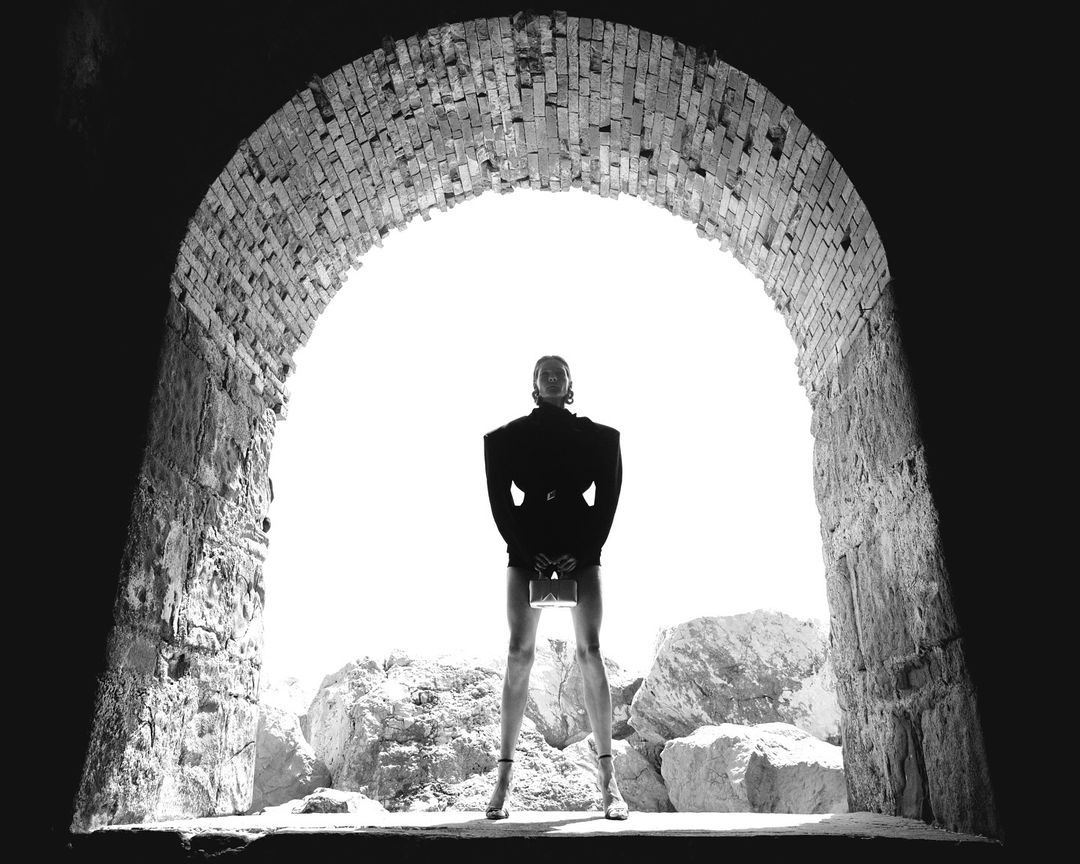 A stylish woman poses in front of a brick archway holding a purse, framed by natural light with a rocky landscape in the background.