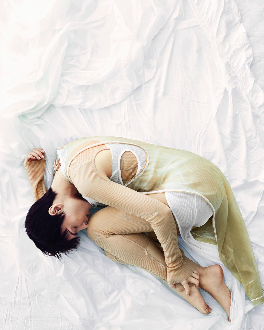 A person is curled on a white textured fabric, wearing a sheer, full-sleeve beige top over a white tank top, with a neutral expression.