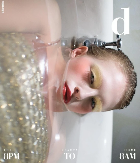 A woman is submerged in a tub with her face visible, adorned in makeup and an embellished top, presenting a high-fashion aesthetic.