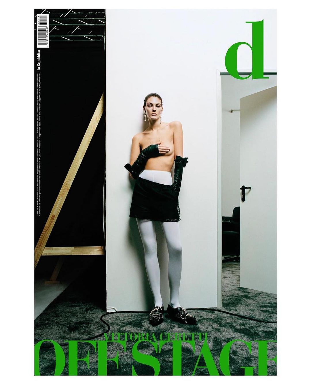 Magazine cover featuring a topless adult female fashion model wearing black gloves, a black skirt, and white tights in a photo studio.