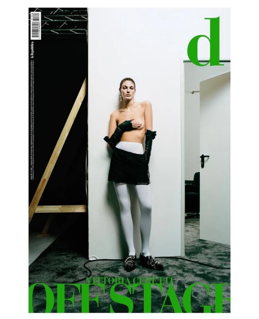 Magazine cover featuring a topless adult female fashion model wearing black gloves, a black skirt, and white tights in a photo studio.