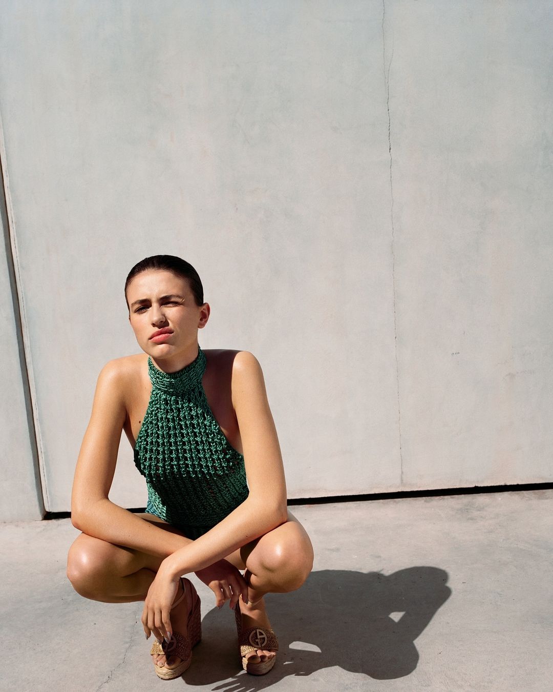 A young adult woman crouches outdoors in a crocheted green top and platform shoes, posing against a plain wall in bright, sunny conditions.