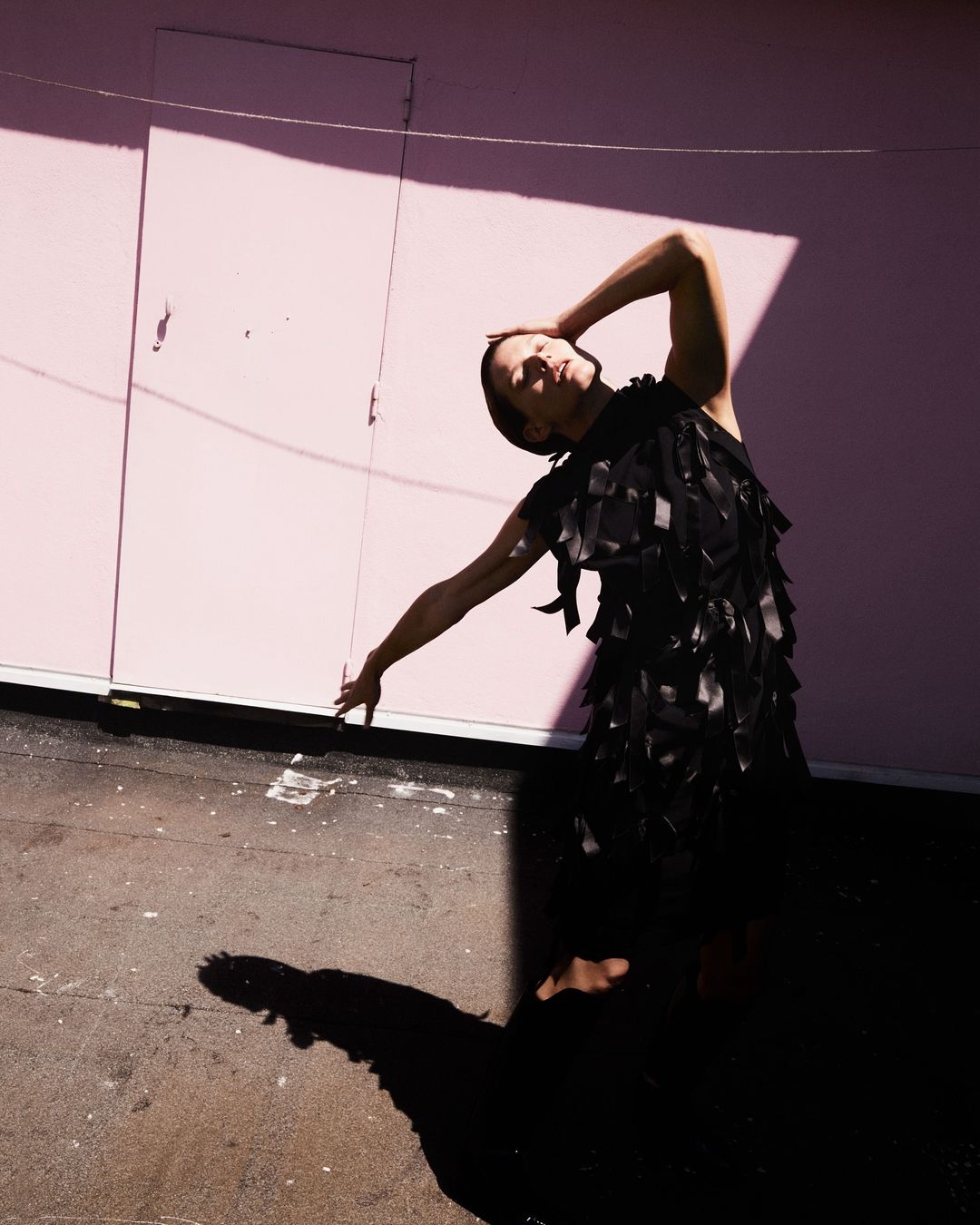Model in a black dress posing against a pink wall with harsh diagonal shadows captured by photographer Vito Fernicola.