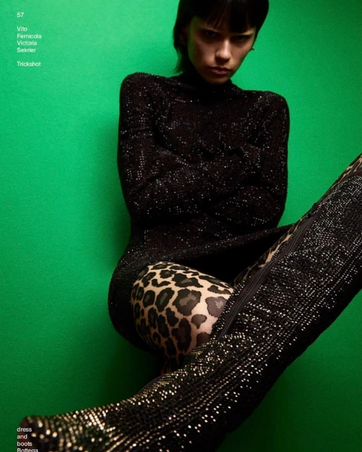 Fashion editorial shot of a woman in a black dress with a leopard print detail and thigh-high boots against a green backdrop.