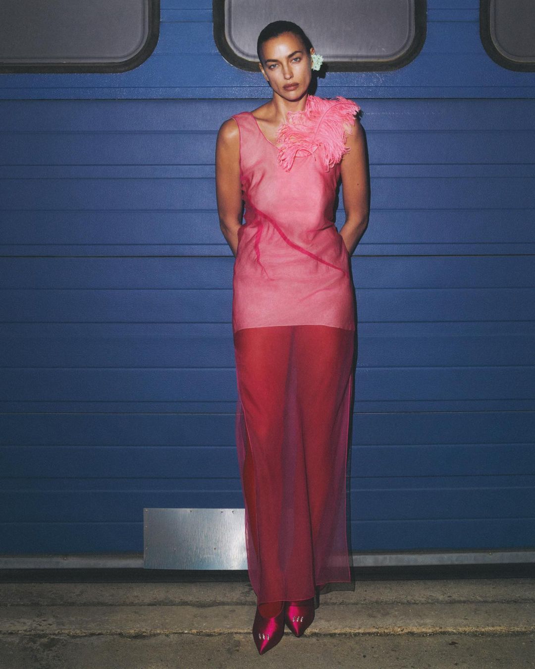 Fashion shot of Irina Shayk in a semi-sheer pink dress against a blue metal wall, showcasing editorial style and beauty.