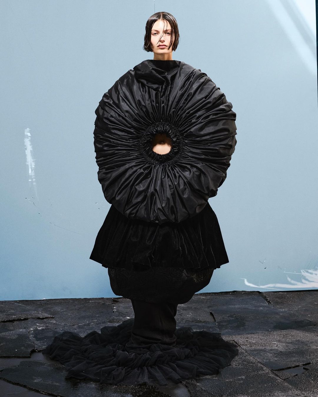Model wearing a voluminous black sculptural dress with a circular gathered bodice against a pale backdrop