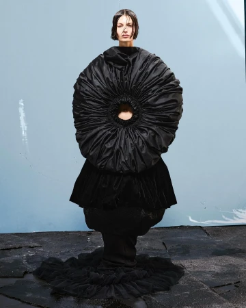 Model wearing a voluminous black sculptural dress with a circular gathered bodice against a pale backdrop