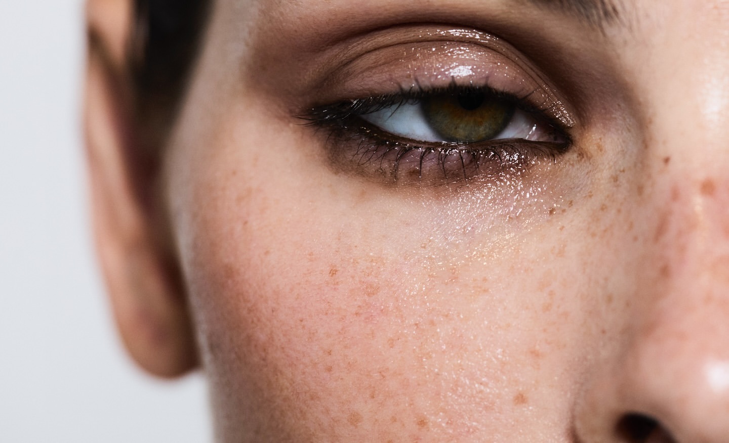 Close-up of a woman's face showing her eye with brown eyeshadow, dark eyeliner, and long lashes. Her skin shows freckles and natural texture.