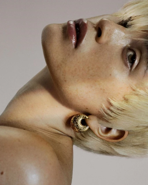 Close-up studio shot of adult female with blonde hair, freckles, and a large, circular, gold earring against a pale pink background.