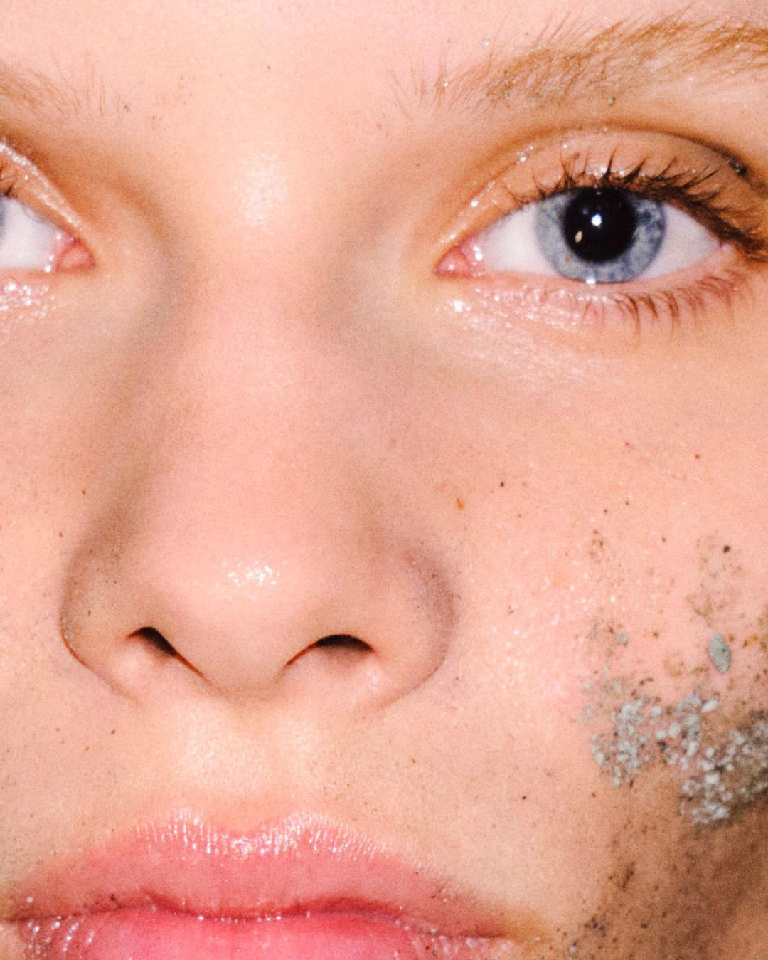 Close-up of a fair-skinned person's face with blue eyes and a neutral expression, skin is adorned with sand or dirt.