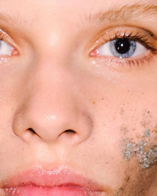 Close-up of a fair-skinned person's face with blue eyes and a neutral expression, skin is adorned with sand or dirt.