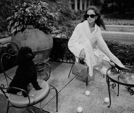 Elegant woman in a white robe lounges on a chair with a black poodle nearby, set against a lush garden backdrop in a black and white shot.