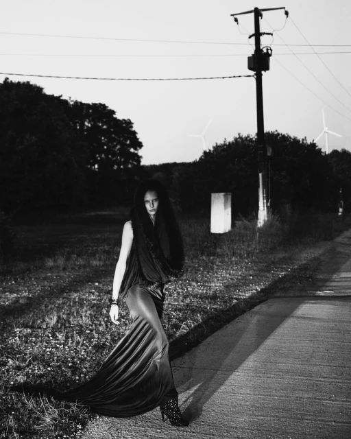 Monochrome outdoor fashion shot featuring a woman in a long gown, posing on a path with windmills and a utility pole in the background.