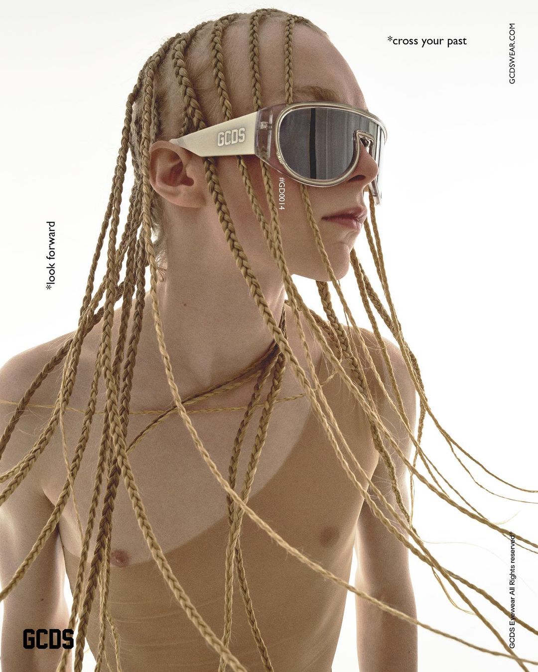 Close-up studio shot of an adult with blonde braided hair, wearing a tan top and translucent sunglasses, against a white backdrop.