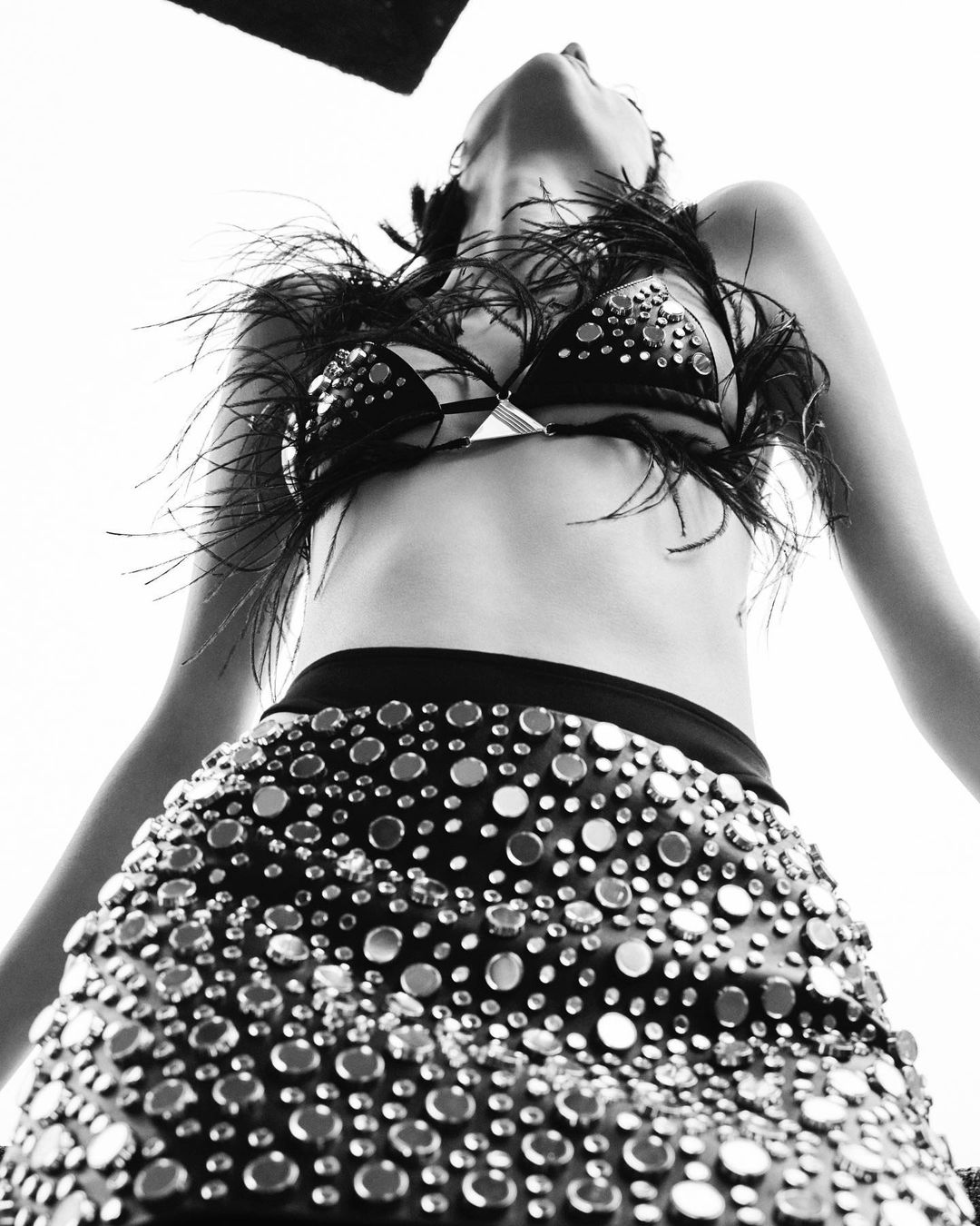 Low-angle shot of a woman in a studded skirt and feather-trimmed bra, looking up into a bright light, emphasizing fashion and style.