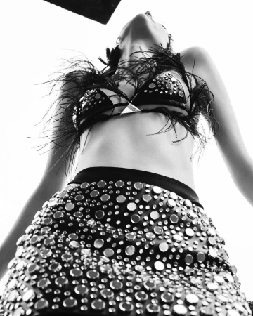 Low-angle shot of a woman in a studded skirt and feather-trimmed bra, looking up into a bright light, emphasizing fashion and style.