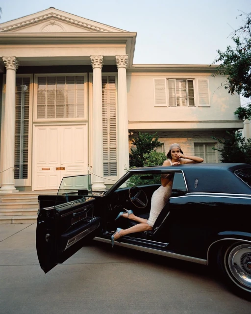 Elegant woman in a beige dress posing beside a vintage black car, with a grand house featuring white columns in the background.