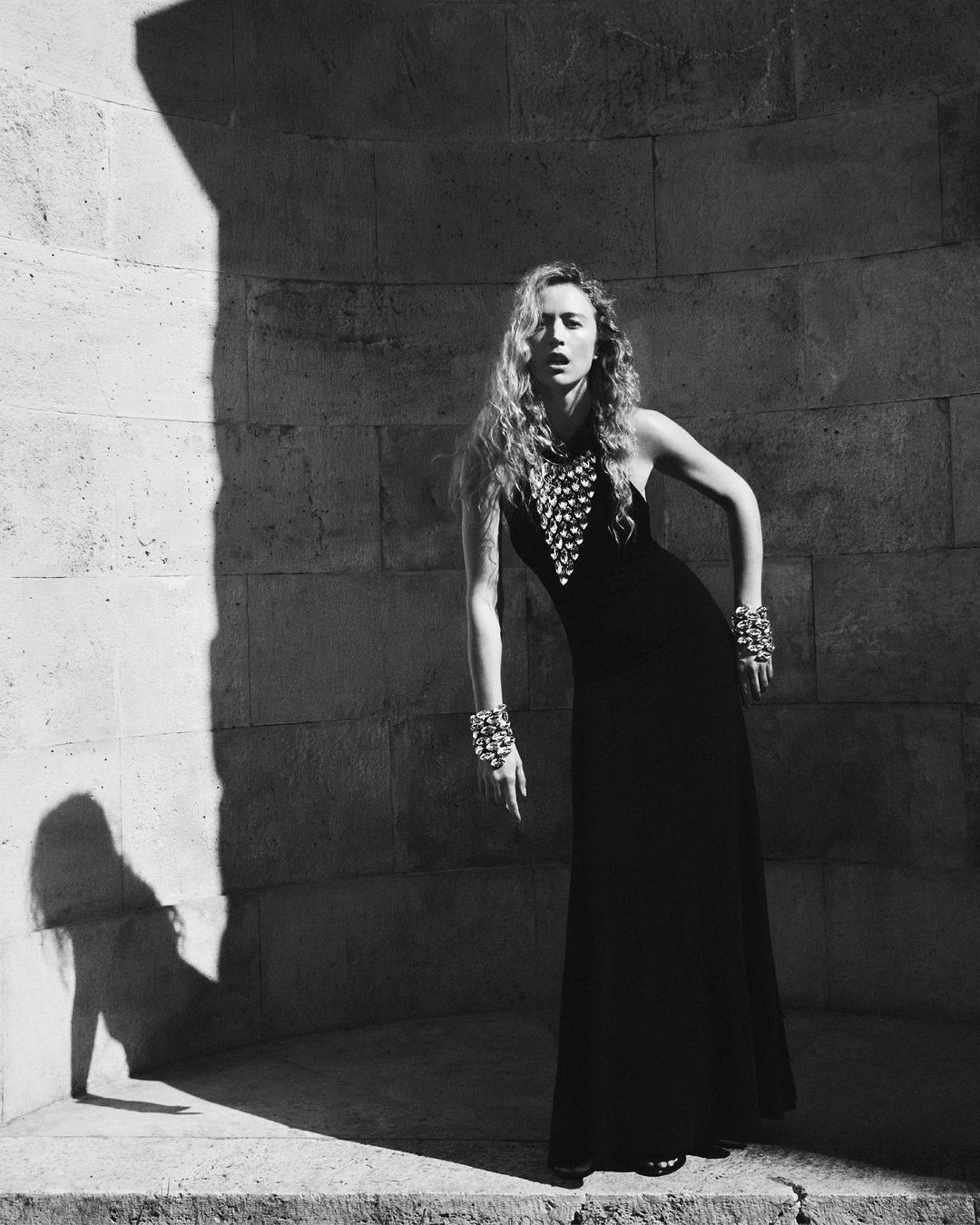 Black and white fashion shot of a woman in a long black gown with studded embellishments, posing in a stone architectural structure.