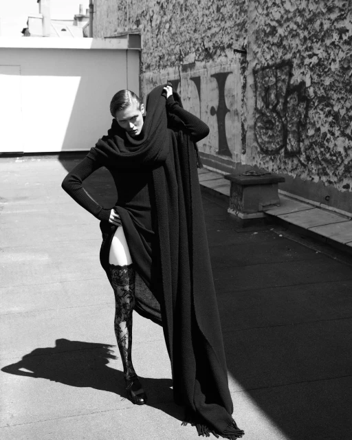 A fashion shot in black and white shows a slender woman in a long, black dress and patterned stockings posing on a rooftop.