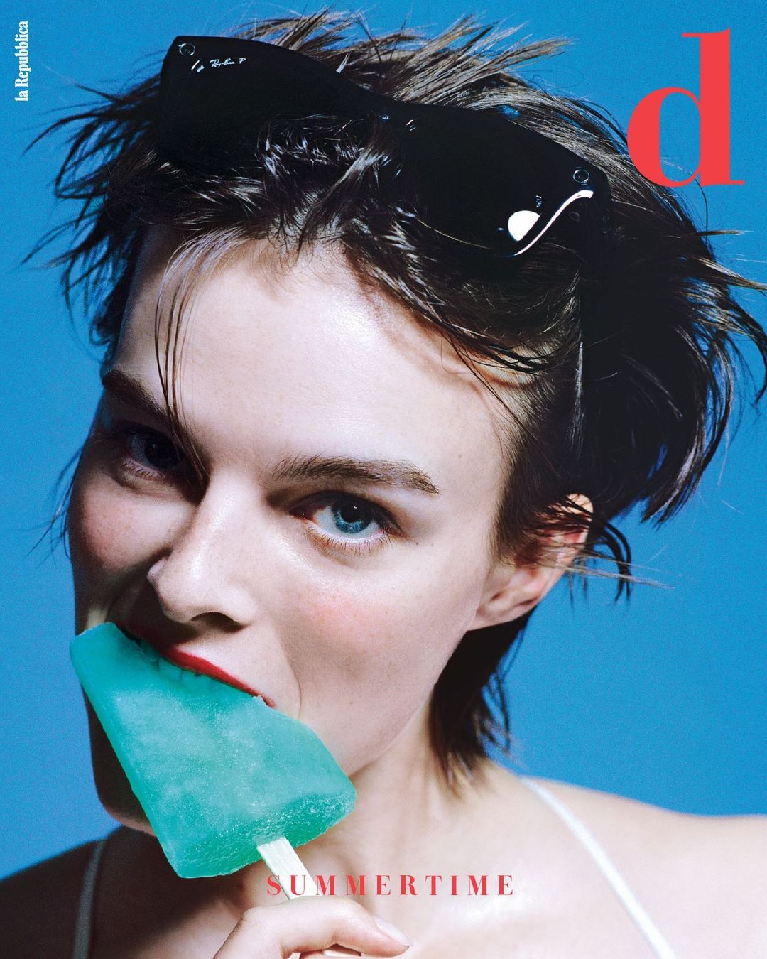 Magazine cover featuring a woman eating a blue popsicle with a blue background, short brown hair, sunglasses, and red lipstick.