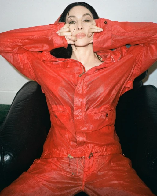 A young woman with dark hair poses in a red jumpsuit against a neutral background, seated in a black chair, styled with slightly damp hair.