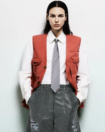 Model in terracotta vest, white shirt, grey tie, and patterned trousers against a plain white background
