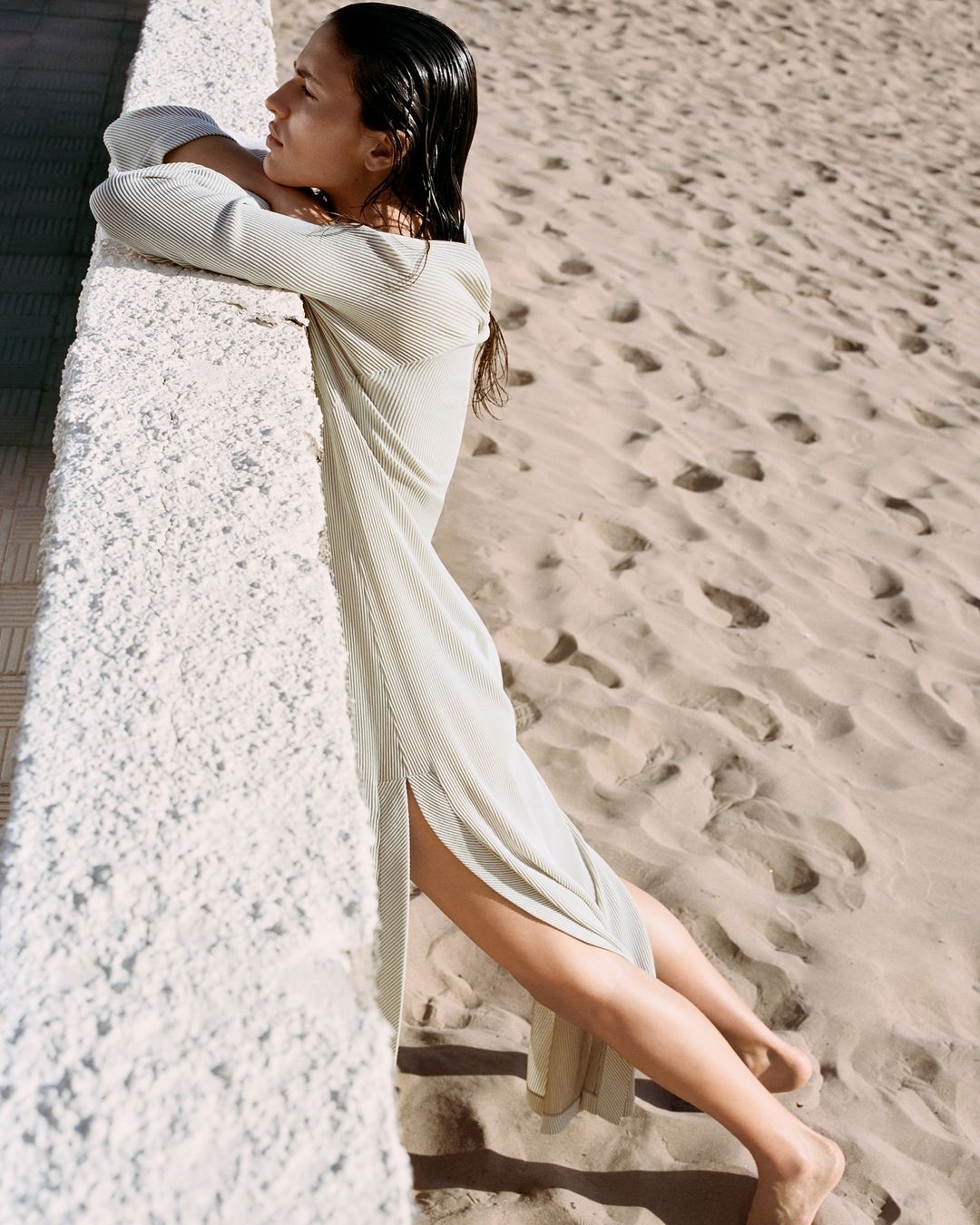 Woman in a knitted maxi dress leans against a stone wall on a sandy beach in a fashion shot with golden hour lighting.
