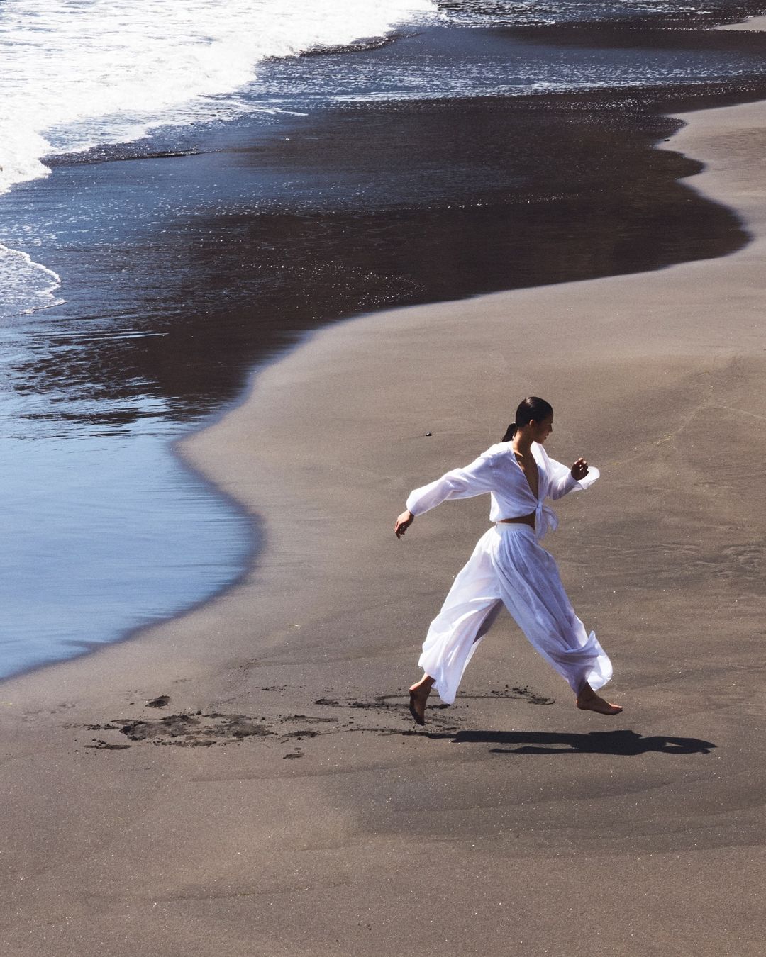 Young woman in white clothes jumps barefoot on a sandy beach, ocean waves behind her in bright daylight. Fashion and fitness.