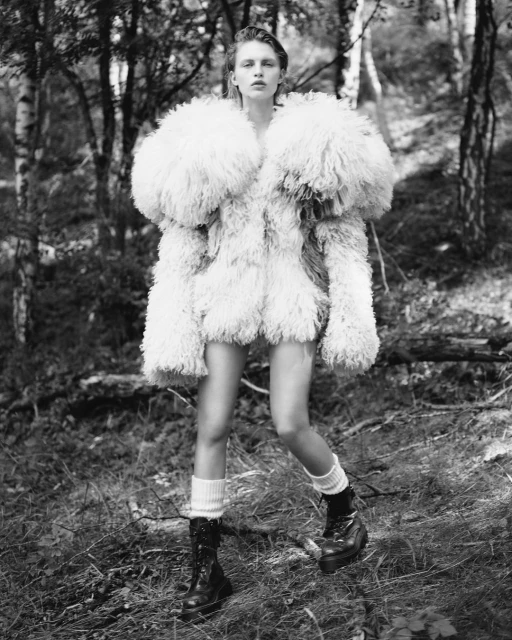 An adult female model is posing in an oversized fur coat, short shorts, socks, and boots in a grayscale woodland setting.