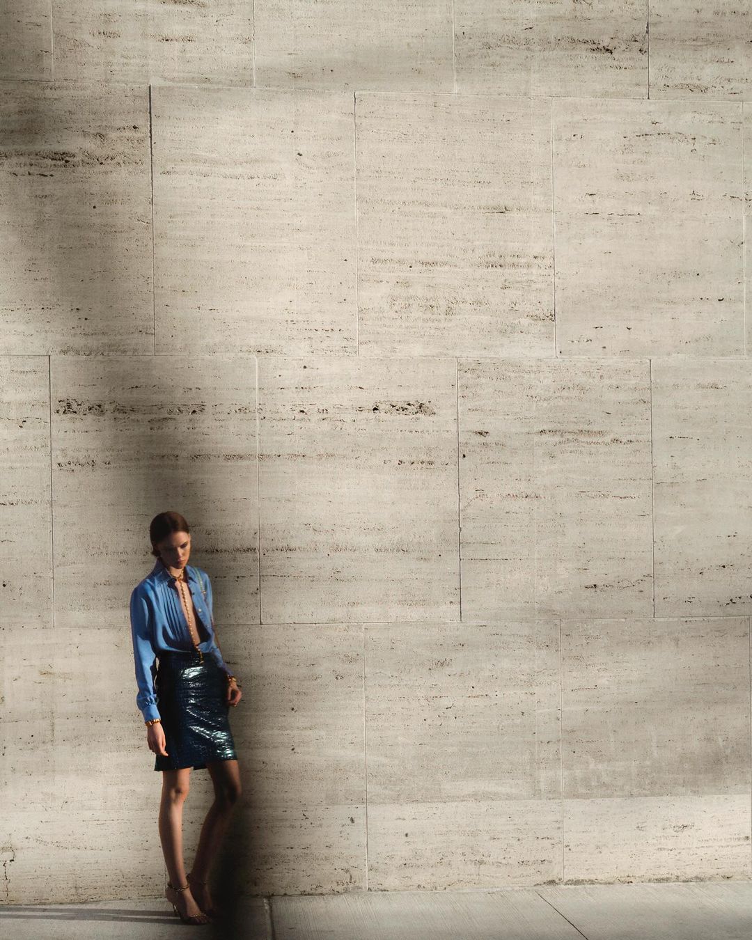 A stylish woman in a blue shirt and short black skirt poses against a textured beige wall in an indoor environment with natural lighting.