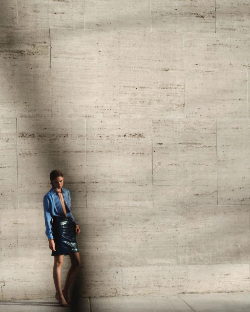 A stylish woman in a blue shirt and short black skirt poses against a textured beige wall in an indoor environment with natural lighting.